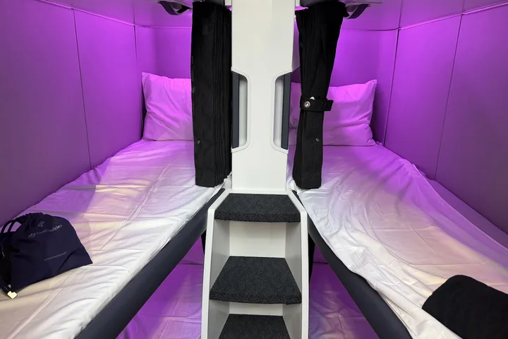 Two side-by-side airplane sleep pods with white bedding and purple ambient lighting, with a ladder between them.