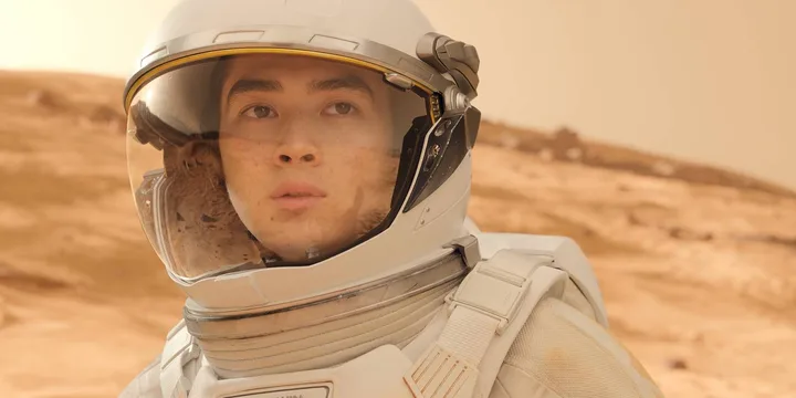 Young man in a white spacesuit helmet on a Mars-like red rocky landscape, serious expression.