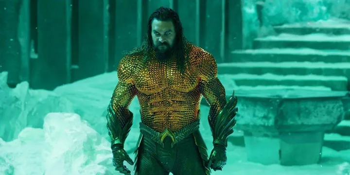 Jason Momoa in gold Aquaman armor stands in a glowing teal underwater setting.