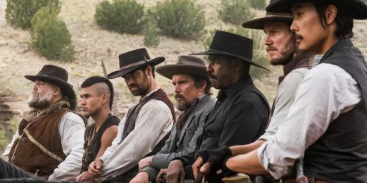 Seven men in cowboy hats and western attire sit together in an outdoor desert setting.