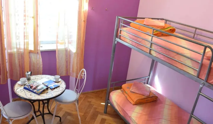Colorful hostel room with metal bunk beds, orange bedding, small bistro table, and purple walls.