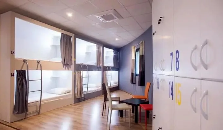 Modern hostel dormitory with pod-style bunk beds, curtains, numbered lockers, and a small table.
