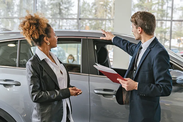 Male car salesman with clipboard gesturing toward a gray car while speaking with a female customer in a dealership.