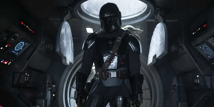 The Mandalorian in dark armor stands inside a spacecraft with Grogu visible on his shoulder.