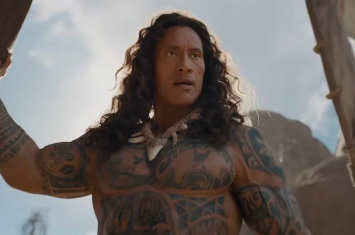 Heavily tattooed muscular man with long curly hair in outdoor fantasy setting, portraying Maui.