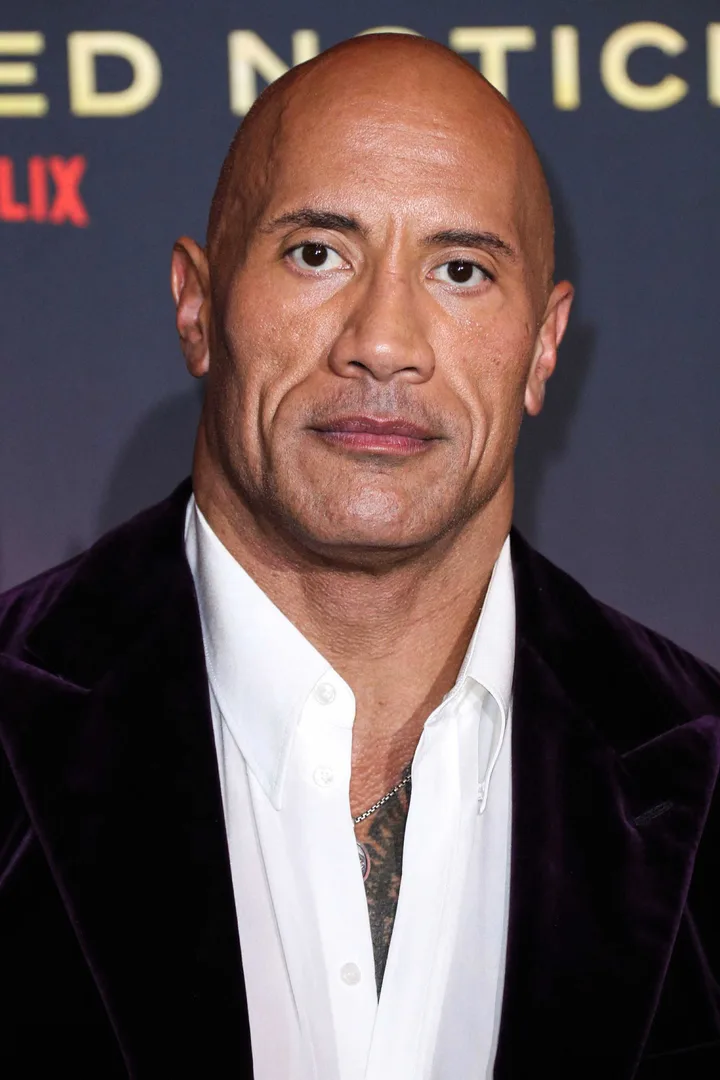 Bald muscular man in purple velvet blazer at a Netflix red carpet event, likely Dwayne Johnson.