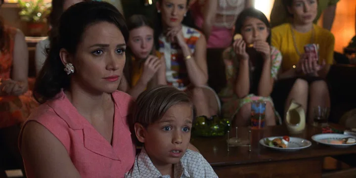 Woman in pink dress holding a young boy, surrounded by children at a retro-styled indoor gathering.