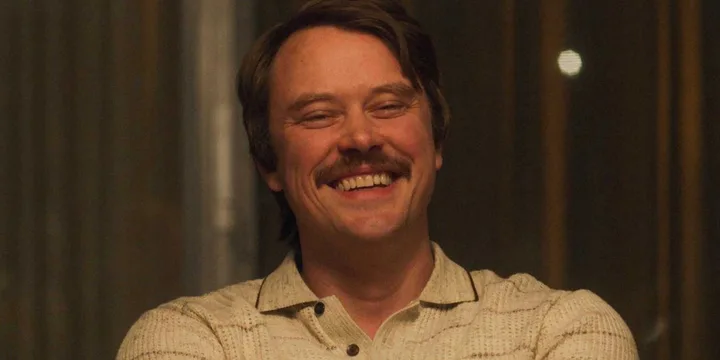 Smiling mustachioed man in a cream knit shirt in a warmly lit, retro interior setting.