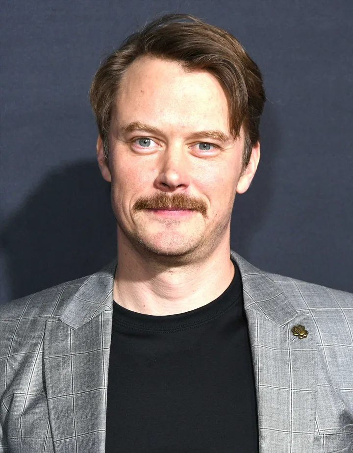 Man with mustache in a grey plaid blazer posing at what appears to be a press or premiere event.