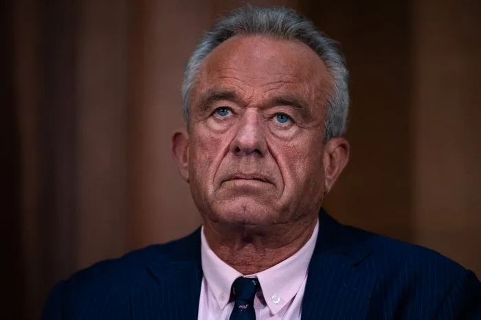 RFK Jr. with a serious expression in a blue suit, looking upward against a warm brown background.