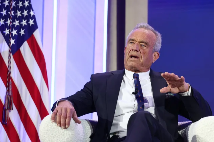 RFK Jr. gesturing while seated next to an American flag at a public event.