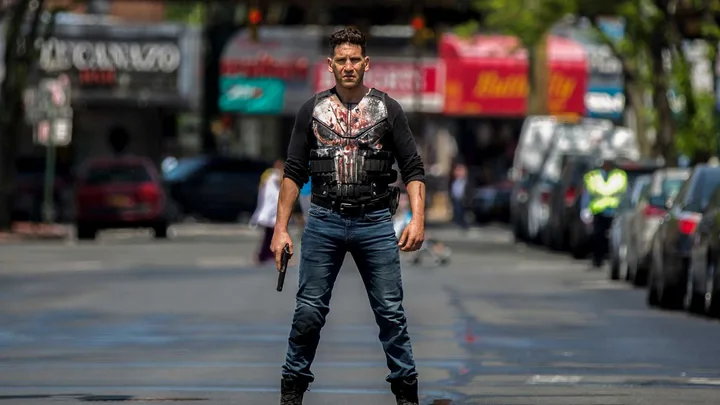 Jon Bernthal as the Punisher in skull vest holding a gun, standing defiantly on a city street.