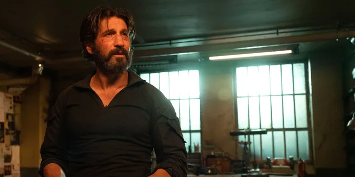 Jon Bernthal as a bearded man in black, standing in a gritty workshop or warehouse setting.