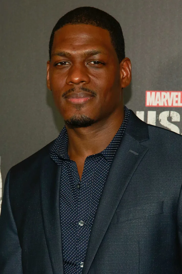 A man in a navy suit at a Marvel's Punisher premiere event, posing against a Marvel-branded backdrop.