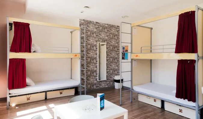 Clean hostel dorm room with metal bunk beds, red curtains, and storage drawers.
