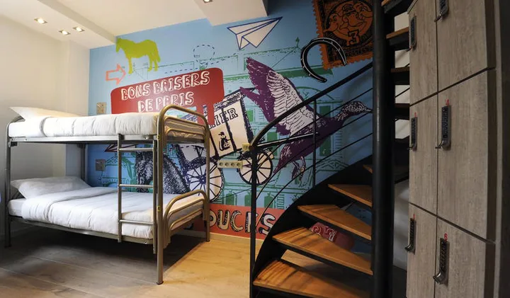 Colorful hostel dorm room with Paris-themed mural, bunk bed, spiral staircase, and lockers.