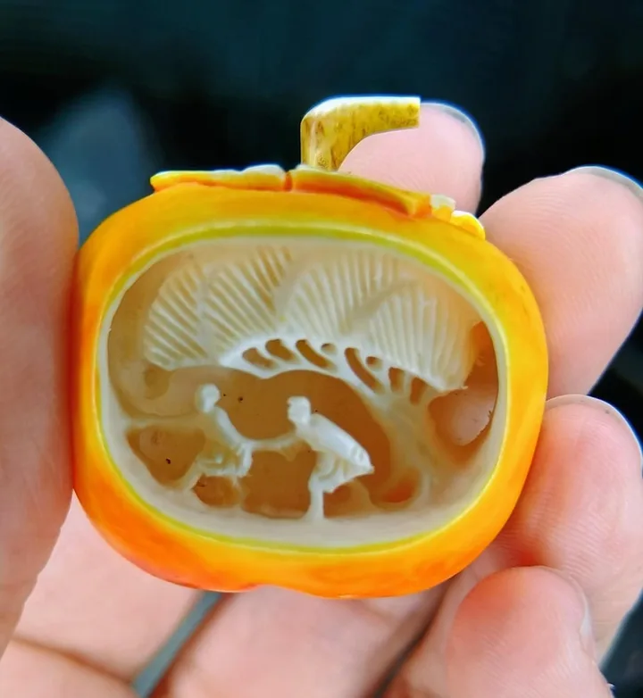 Hand holding a small antique cameo brooch carved with two figures under a tree, orange shell frame.