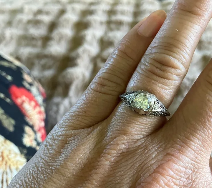 An antique filigree diamond engagement ring worn on a finger against a floral fabric background.