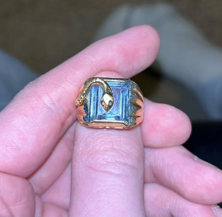 Hand holding an ornate vintage gold ring with a large blue aquamarine stone and snake-motif shoulders.
