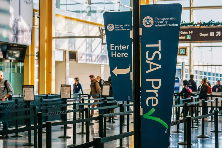 TSA PreCheck lane entrance signs at a bright, busy airport security area with travelers queuing.
