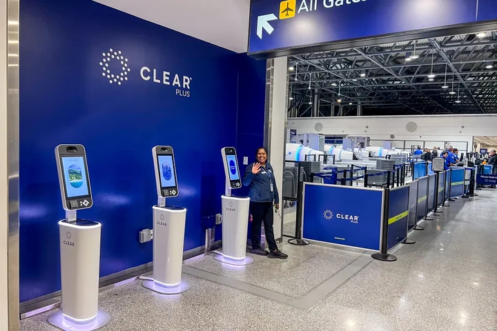 CLEAR Plus biometric kiosks at an airport with a staff member waving beside them.
