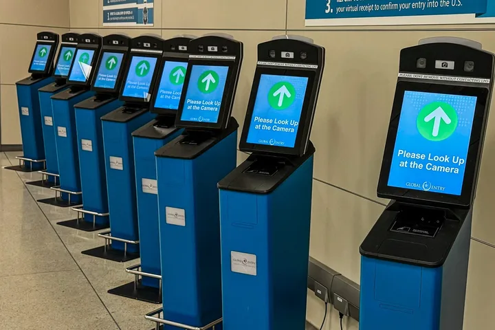 Row of Global Entry self-service kiosks with facial recognition screens in an airport arrivals hall.