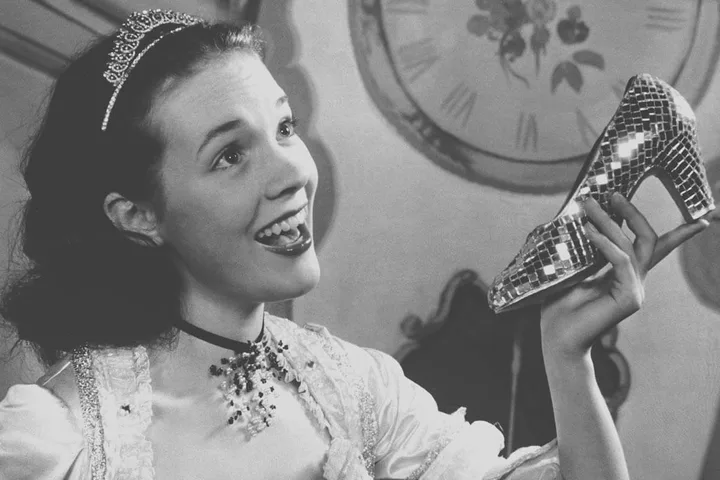 Black and white vintage photo of a smiling young woman in period costume holding a glittery glass slipper.
