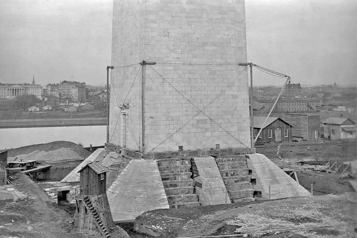 Historical black-and-white photo of Washington Monument base under construction with scaffolding and city behind.