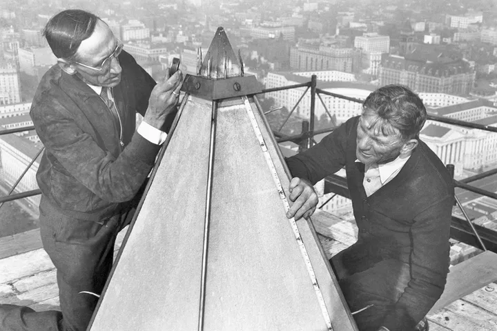 Two workers examining the aluminum apex of the Washington Monument atop the structure with city below.