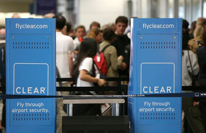 Two blue CLEAR kiosks reading 'Fly through airport security' with travelers lined up behind them.