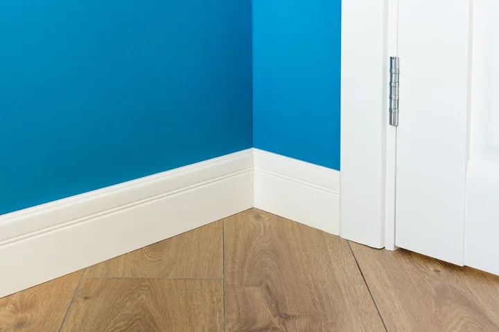 White baseboard molding meeting blue wall and hardwood floor in room corner with door.