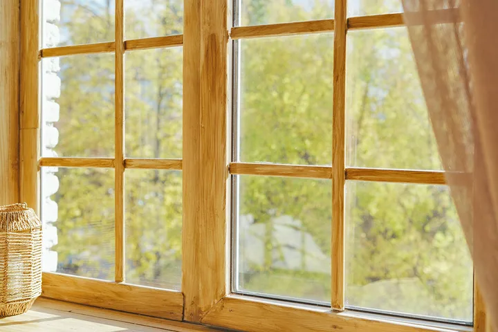 Rustic wooden-framed window with multi-pane grid overlooking green trees with sheer curtain.