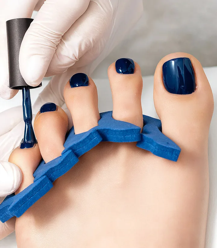 Gloved technician applying navy blue nail polish to toes held apart by a foam toe separator.