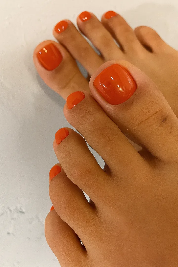 Close-up of two feet with glossy orange pedicure against a white surface.
