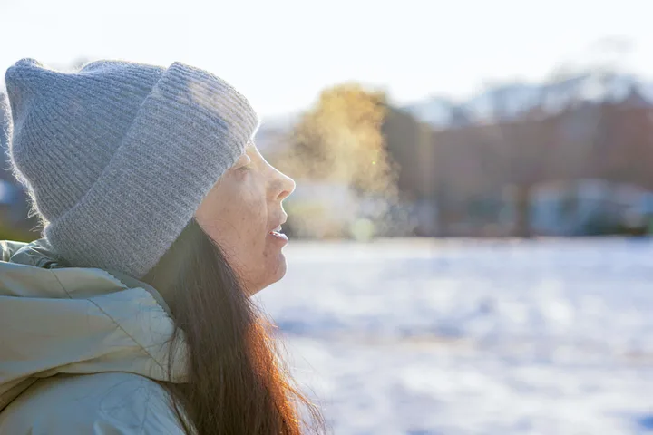 Woman in gray beanie exhaling visible breath vapor in cold snowy outdoor setting.