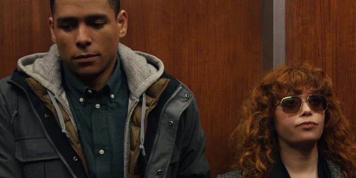 A man and a red-haired woman wearing sunglasses stand together in a wood-paneled elevator, looking subdued.