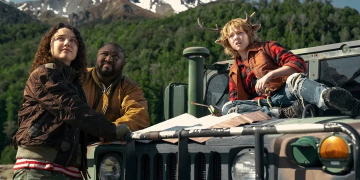 Three characters including a child with antlers look upward while leaning on a military vehicle with mountains behind th