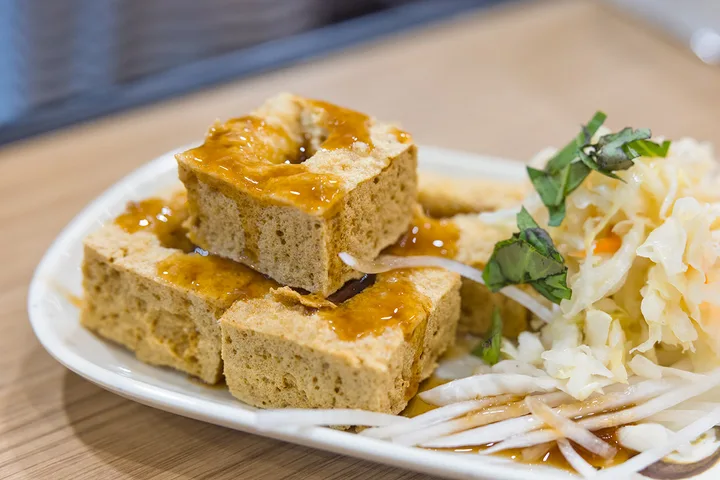 Plate of stinky tofu cubes with brown sauce and pickled cabbage garnish on a wooden table.