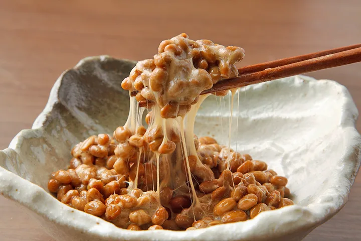 Chopsticks lifting sticky, stringy natto (fermented soybeans) from a ceramic bowl.