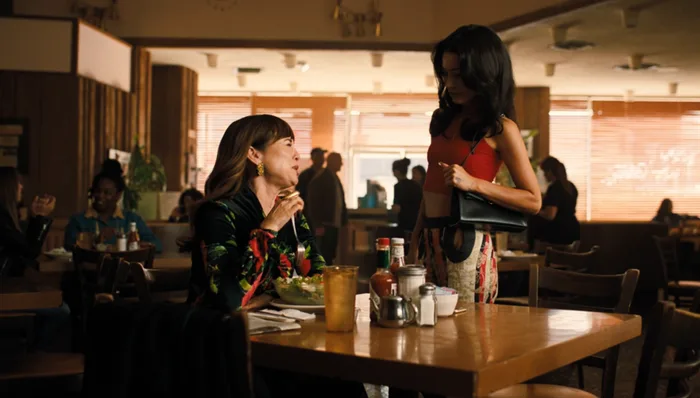 Two women in a retro diner, one seated eating and one standing holding a bag, TV scene.