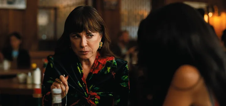 Older woman in a floral dress speaking intensely across a table in a warmly lit diner, TV scene.