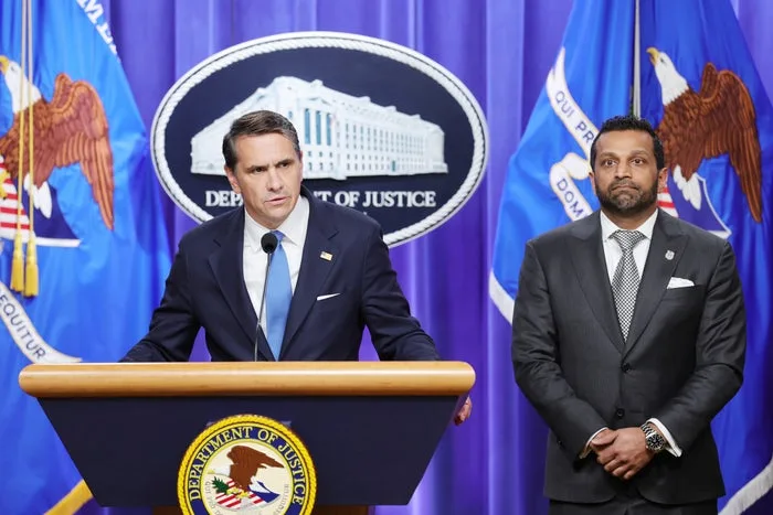 Two men in suits at a Department of Justice press conference podium with DOJ flags.
