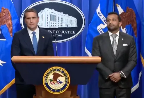 Two men in suits at a Department of Justice press conference, wide shot with DOJ seal visible.