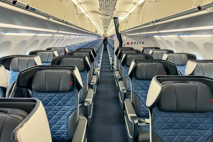 Empty Delta first class cabin aisle view with quilted blue seats and Delta logo visible at the front.