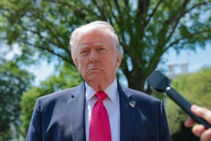 Older man in blue suit and red tie speaking outdoors near a microphone, looking serious.