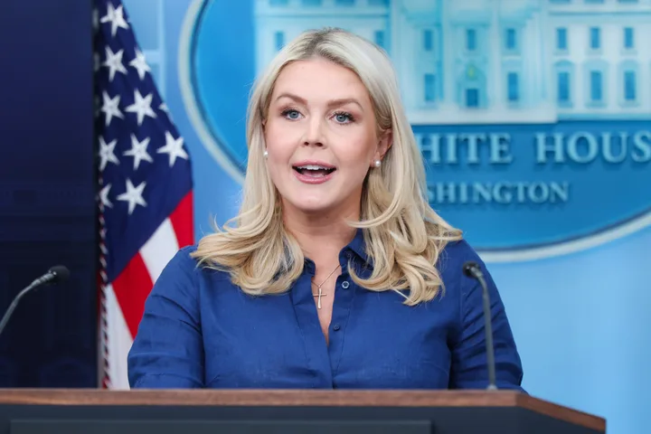 Blonde woman in blue shirt speaking at White House press briefing podium, animated expression.