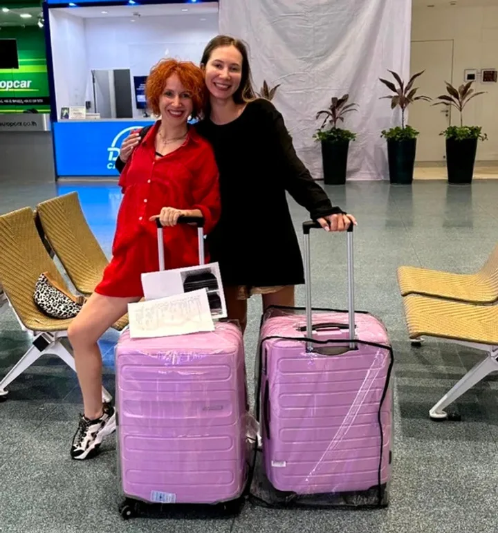 Two smiling women posing with matching pink wrapped suitcases in an airport terminal.
