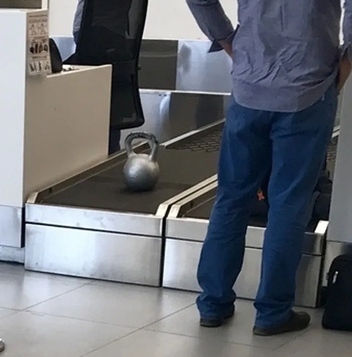 A kettlebell sitting alone on an airport security baggage conveyor belt next to a standing passenger.