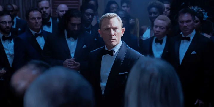 Man in tuxedo stands stoically amid a crowd of formally dressed people in dim lighting.