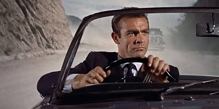 Man in suit driving a convertible car along a dusty road in a vintage film still.